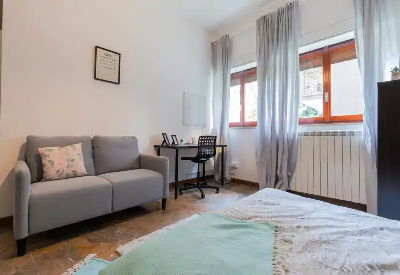 Dormitorio luminoso y ordenado con cama en primer plano, un escritorio junto a la ventana y un sofá de dos plazas acogedor — ideal para una persona o pareja.