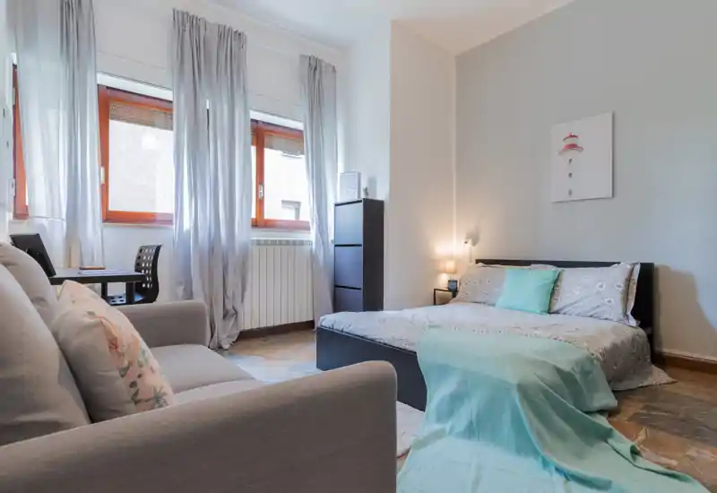 Dormitorio luminoso y bien presentado con cama doble, manta y cojines turquesa, amplias ventanas que aportan luz natural y una pequeña zona de estar — perfecto para un anuncio de alquiler.