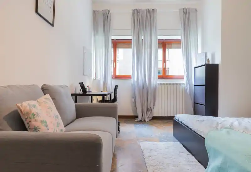 Dormitorio luminoso de estilo sencillo con cama doble visible, pequeño escritorio junto a la ventana y un sofá cómodo — ideal para una persona o pareja. La luz natural entra por la gran ventana y los textiles suaves crean un ambiente acogedor.