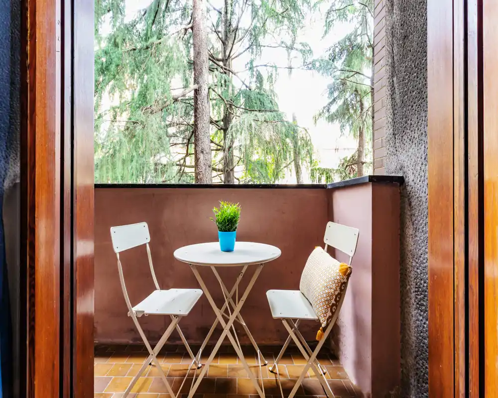 Small furnished balcony with a round table, two folding chairs and a decorative cushion; pleasant greenery visible beyond the railing, ideal for morning coffee.