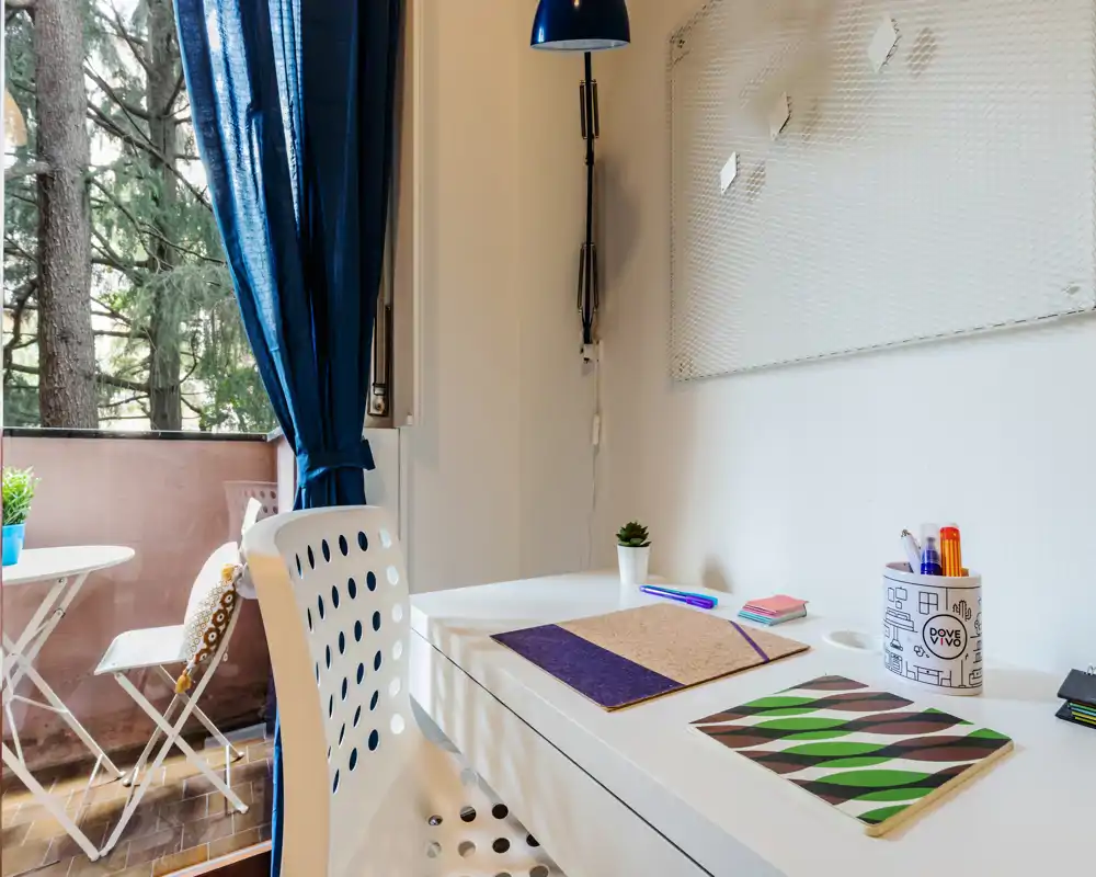Bright, tidy living/work corner with a white desk, chair and a small balcony visible through a blue-curtained window. The space is styled with stationery, a small plant and soft accents, suitable for remote work or study.