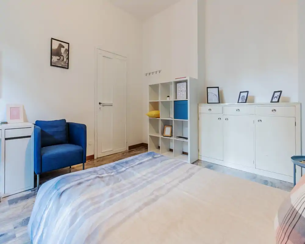 Bright, neatly styled bedroom with a large visible bed in the foreground, a blue armchair, shelving unit and white storage cabinet—clean, airy and inviting.