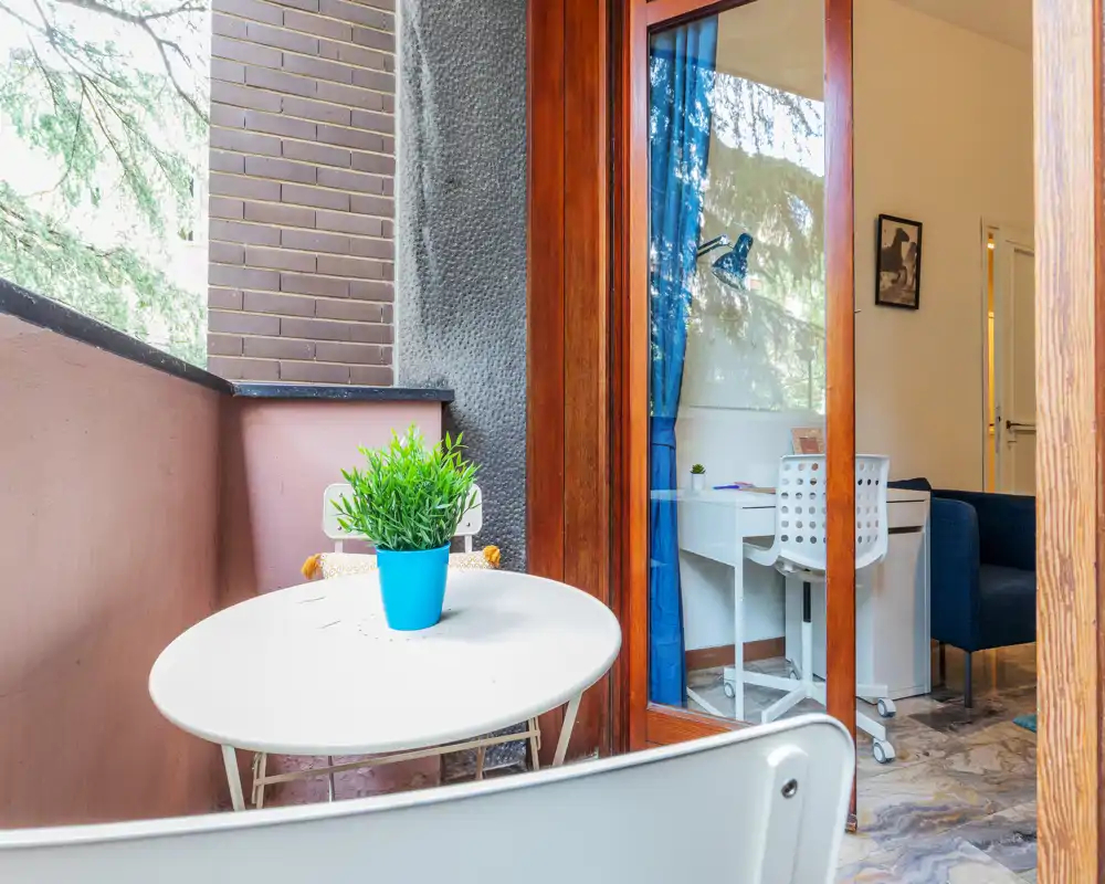 Small private balcony with a round table and chairs, a potted plant as centerpiece, and a sliding glass door leading into a bright interior workspace.