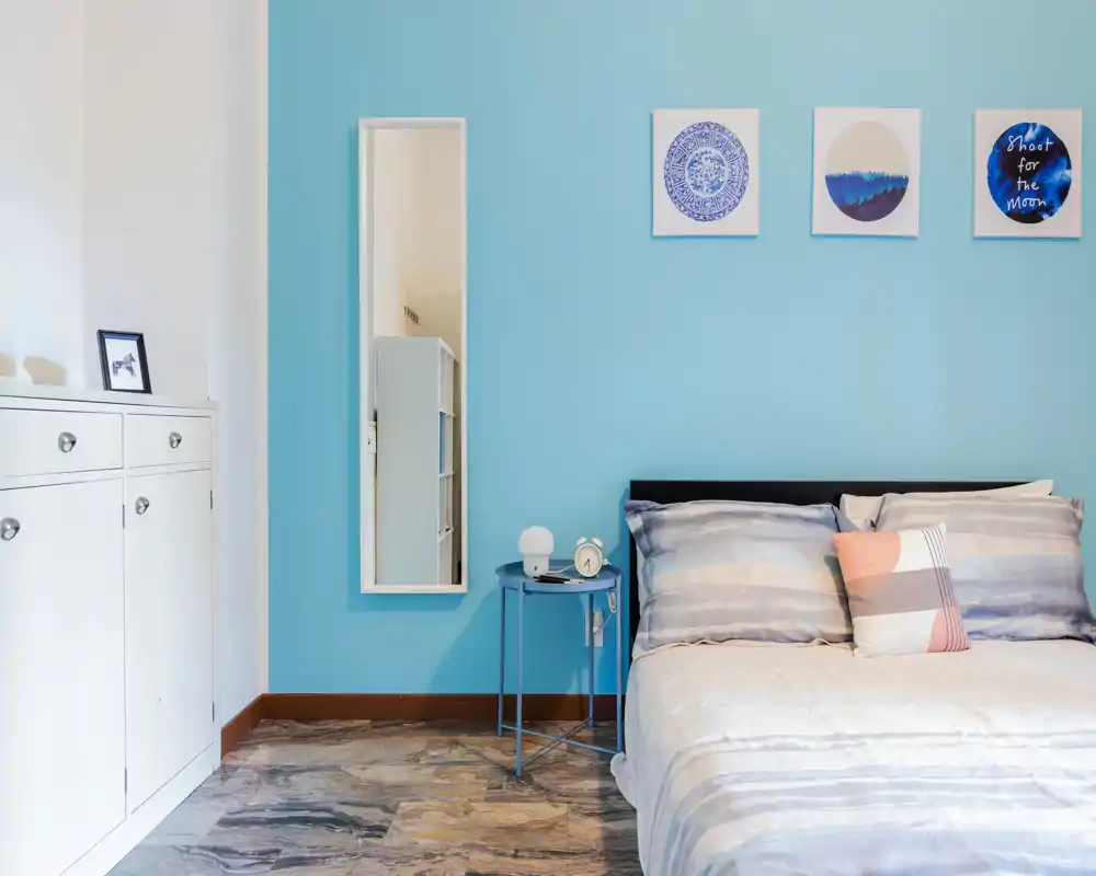 Bright, modern bedroom with a neatly made double bed against a blue accent wall, bedside table with lamp and alarm clock, and tasteful wall art.