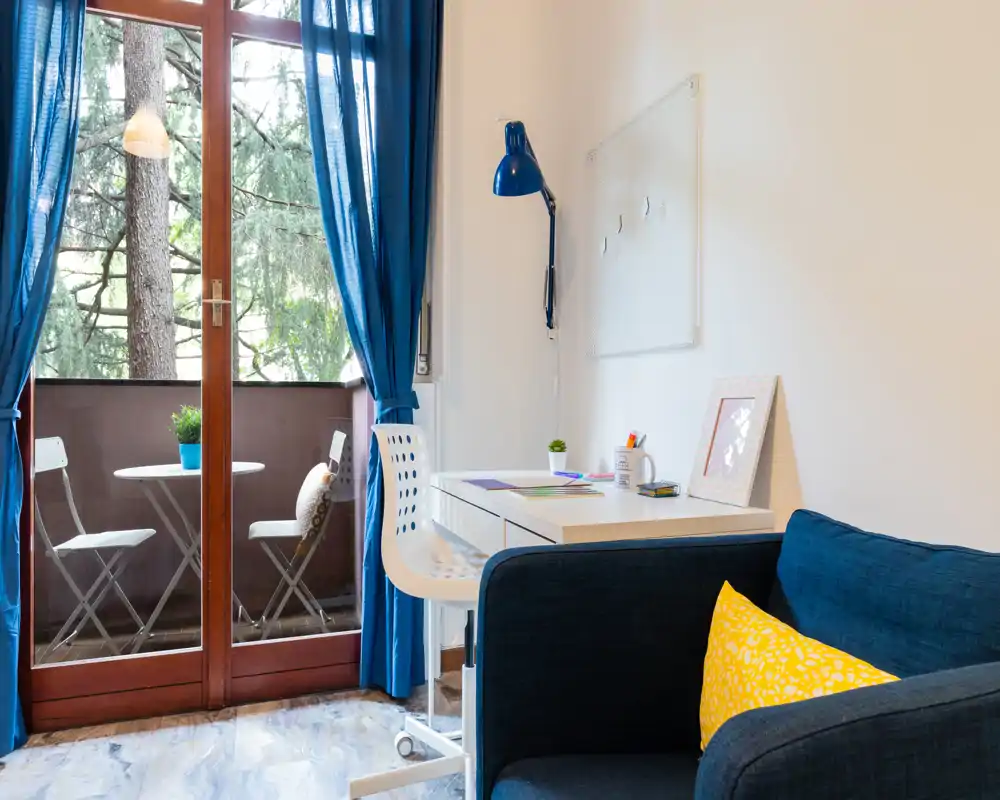 Bright, compact living area with a small balcony: a navy sofa with a yellow cushion, a white desk and chair, and glass doors opening to a bistro table outside.