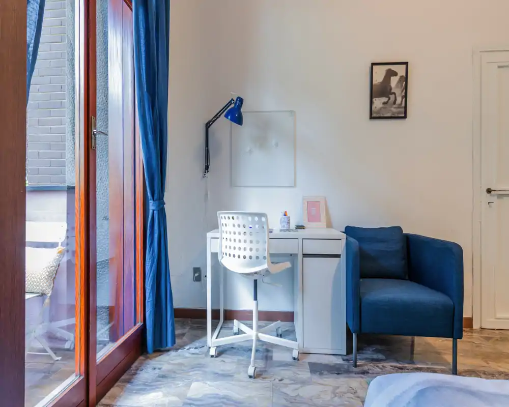 Bright bedroom corner with a small white desk, study lamp and a blue armchair next to a glass door leading to a balcony; a portion of the bed is visible in the foreground.