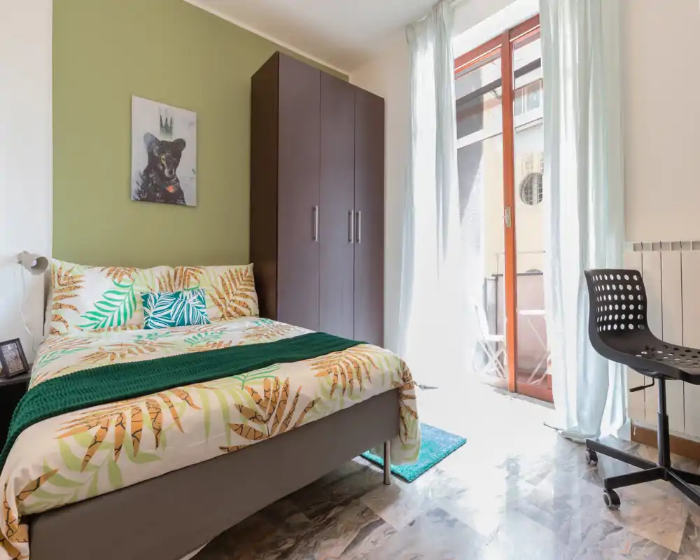 Chambre lumineuse et accueillante avec un lit double aux draps à motifs feuilles, une grande armoire et une porte-fenêtre laissant entrer la lumière du jour.