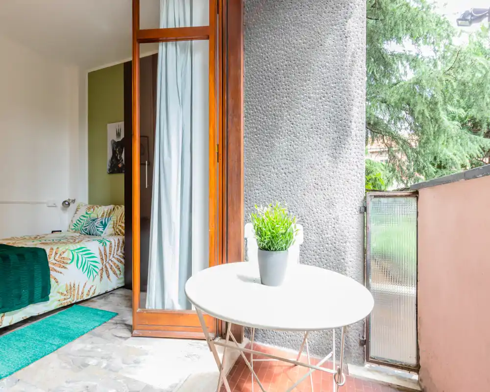 Chambre lumineuse avec une porte coulissante donnant sur un petit balcon. Le lit est habillé de linge à motif tropical et d'un tapis vert ; la pièce bénéficie d'une abondante lumière naturelle.