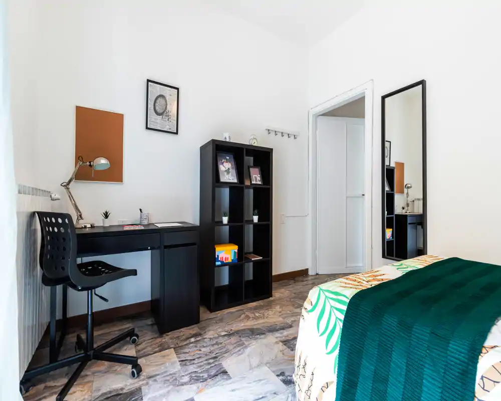 Chambre lumineuse et moderne avec lit bien fait, plaid vert et couette à motif, équipée d'un bureau et d'une étagère — idéale pour un étudiant ou une personne seule.
