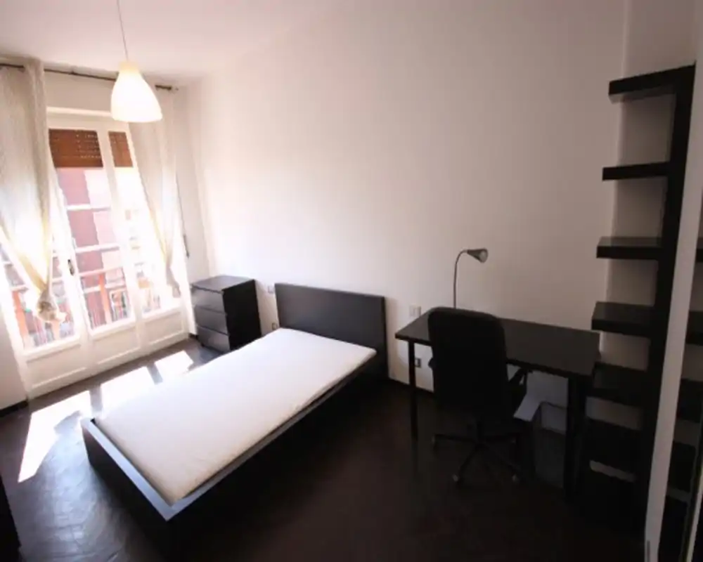 Dormitorio luminoso y minimalista con cama baja, escritorio y estanterías. La gran ventana ofrece mucha luz natural y una sensación limpia y ordenada, ideal para estudiante o persona sola.