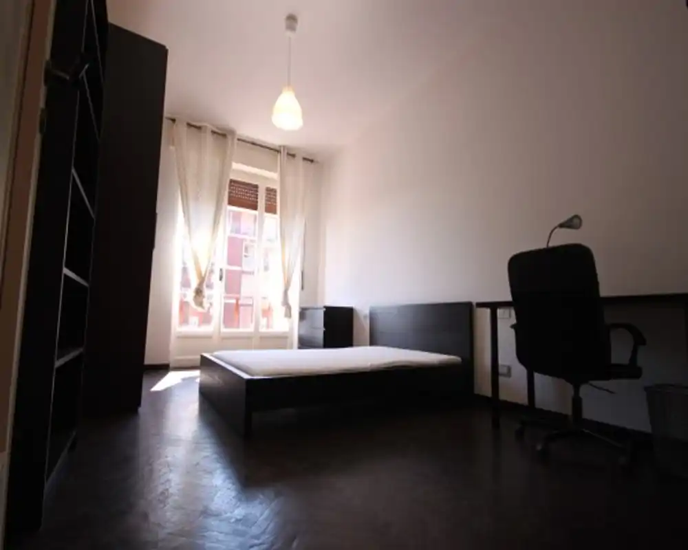 Dormitorio luminoso y minimalista con cama baja, escritorio y estantería; una gran ventana con cortinas aporta abundante luz natural.