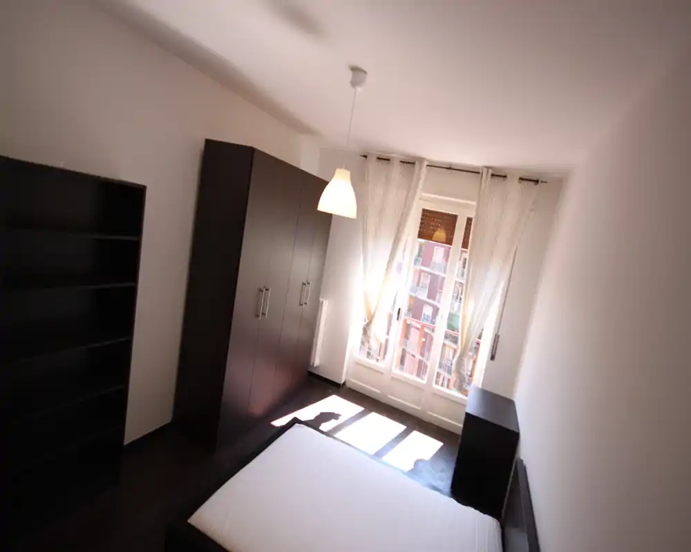 Dormitorio luminoso con cama doble, armario oscuro y estantería, gran ventana con cortinas translúcidas que filtran la luz natural. Mobiliario limpio y minimalista, atractivo para anuncios de alquiler.