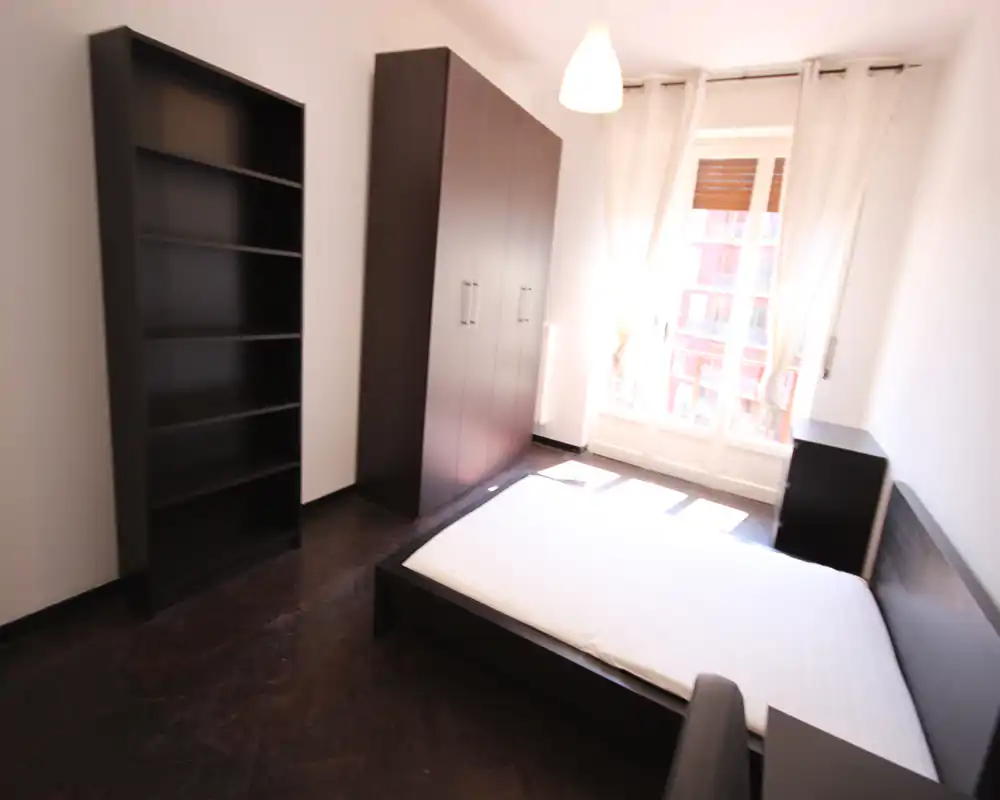 Dormitorio luminoso y amueblado de forma sencilla con cama doble, armario y estantería; gran ventana con cortinas que aporta mucha luz natural.