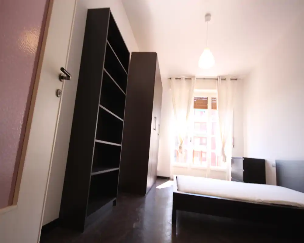 Dormitorio luminoso con cama doble sencilla, gran armario y estanterías; amplia ventana con cortinas translúcidas que aporta mucha luz natural y un aspecto minimalista.