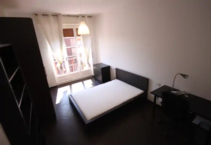 Dormitorio luminoso y minimalista con cama doble, escritorio y armario; gran ventana con luz natural y vista al balcón.