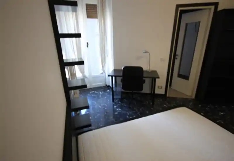 Dormitorio luminoso y sencillo con un colchón grande en primer plano, un escritorio junto a la ventana y estanterías minimalistas — adecuado para una persona o estancias cortas.