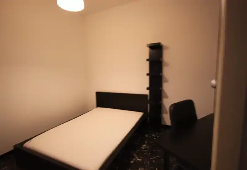 Dormitorio sencillo amueblado con cama individual, colchón y una estantería estrecha; luz de techo cálida y un escritorio pequeño crean un espacio para dormir compacto y funcional.