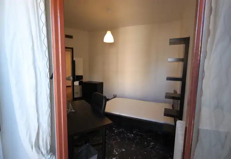 Camera singola luminosa con letto moderno, scrivania e mensole. Luce naturale dalla finestra e arredi semplici e funzionali, ideale per uno studente o un occupante singolo.