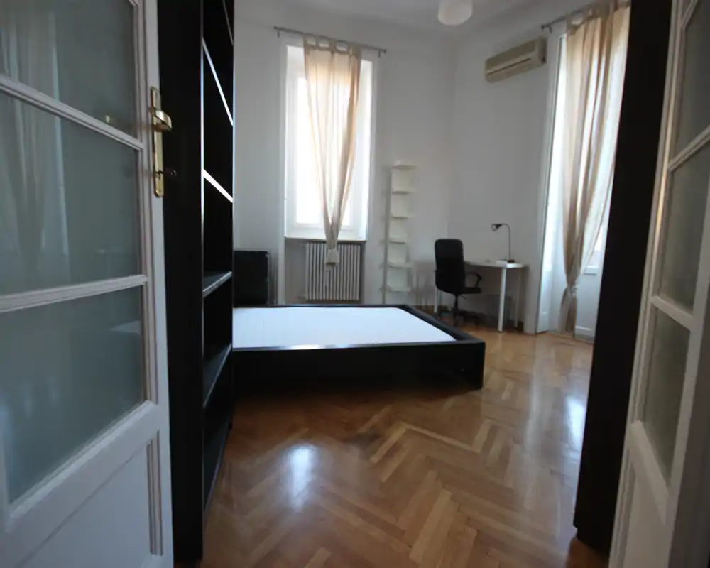 Dormitorio amplio y luminoso con una cama de perfil bajo, grandes ventanas con cortinas ligeras, suelo de parquet y un pequeño escritorio ideal para trabajar.