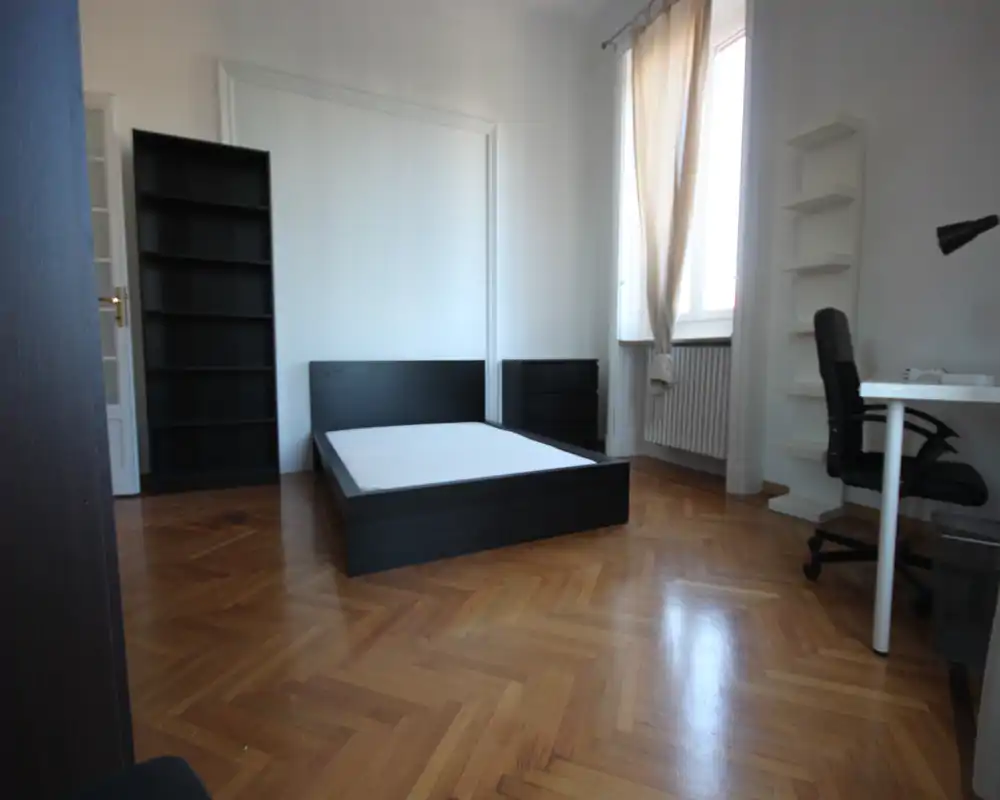 Dormitorio luminoso y minimalista con una base de cama baja oscura, colchón, estantería y escritorio junto a la ventana. Gran ventana con cortinas que aporta luz natural y el parquet pulido da calidez al espacio.