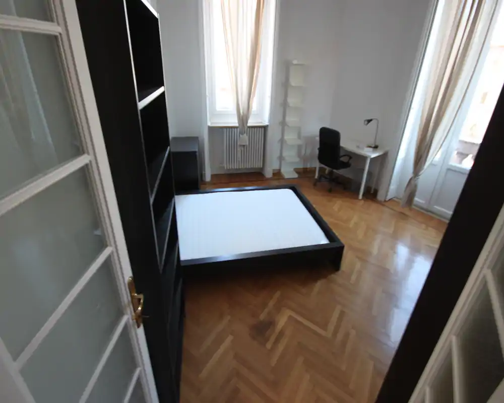 Dormitorio luminoso y minimalista con cama baja en plataforma, suelo de parquet, escritorio y luz natural de dos ventanas.