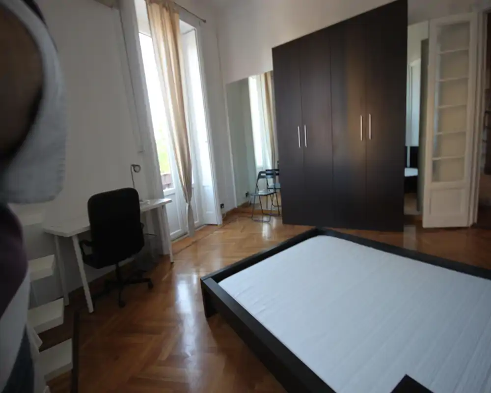 Dormitorio luminoso y espacioso con cama baja, gran armario con paneles espejo, suelo de parqué y escritorio junto a la ventana que aporta luz natural.