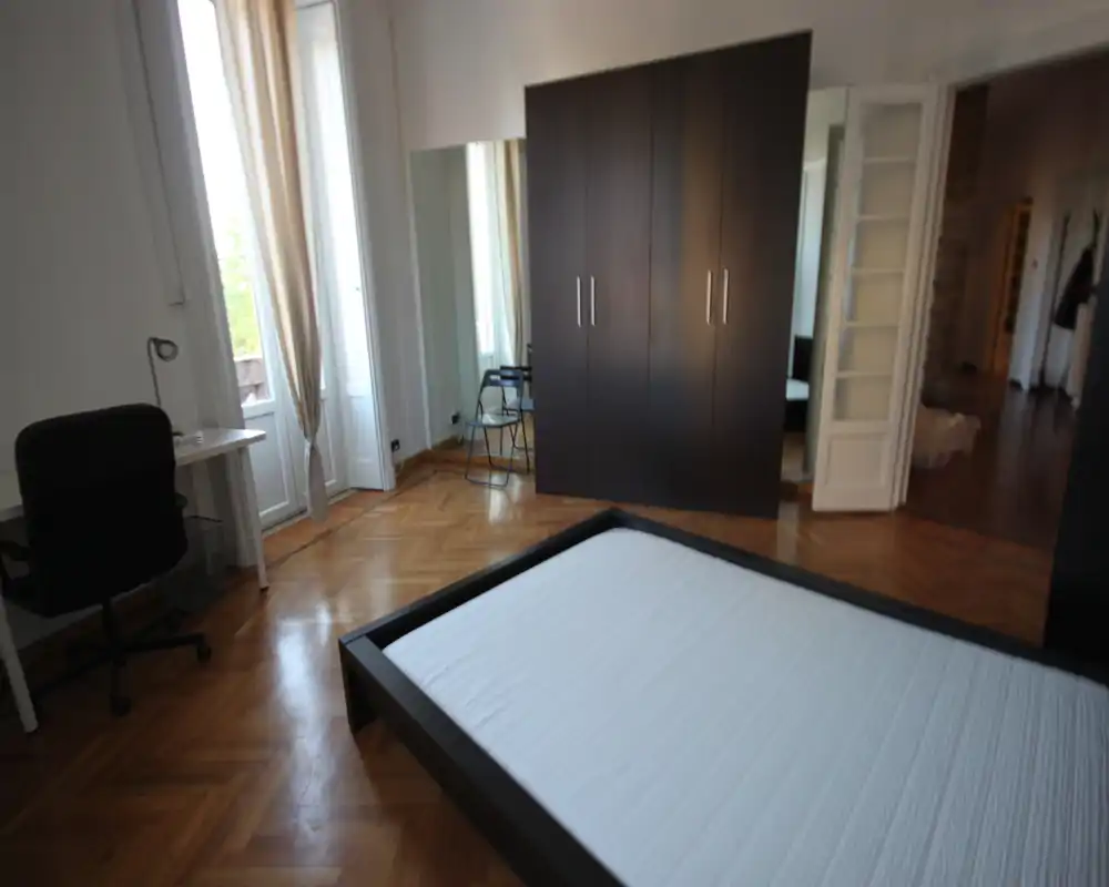 Dormitorio luminoso y amplio con una cama baja moderna, armario grande y ventanas altas que llenan la habitación de luz natural.