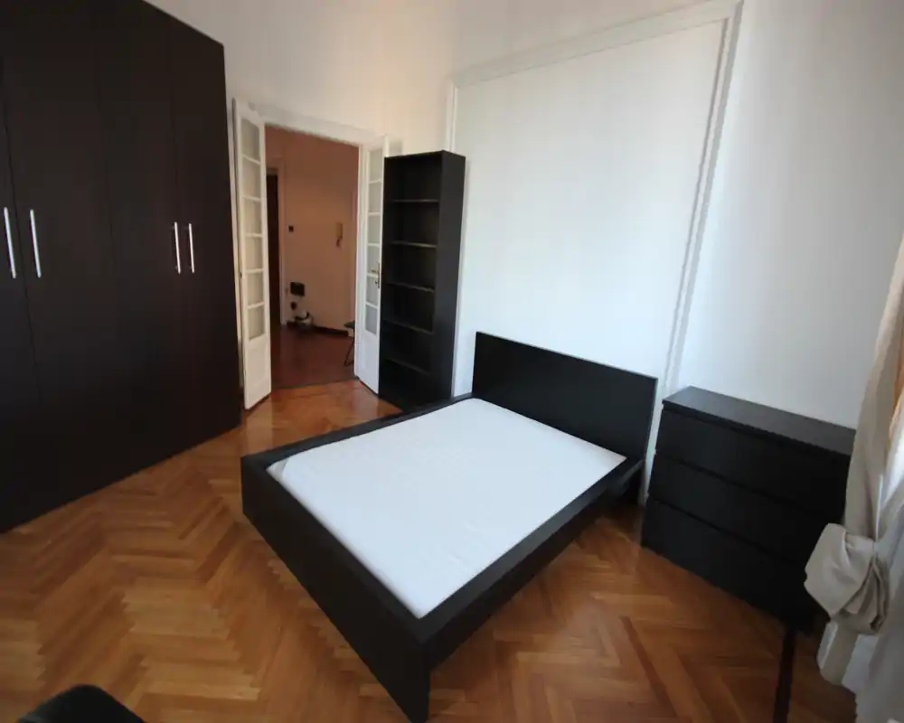 Dormitorio luminoso y minimalista con una amplia estructura de cama en madera oscura, armario y cómoda a juego, y suelo de parquet: espacio de descanso limpio y contemporáneo con luz natural.