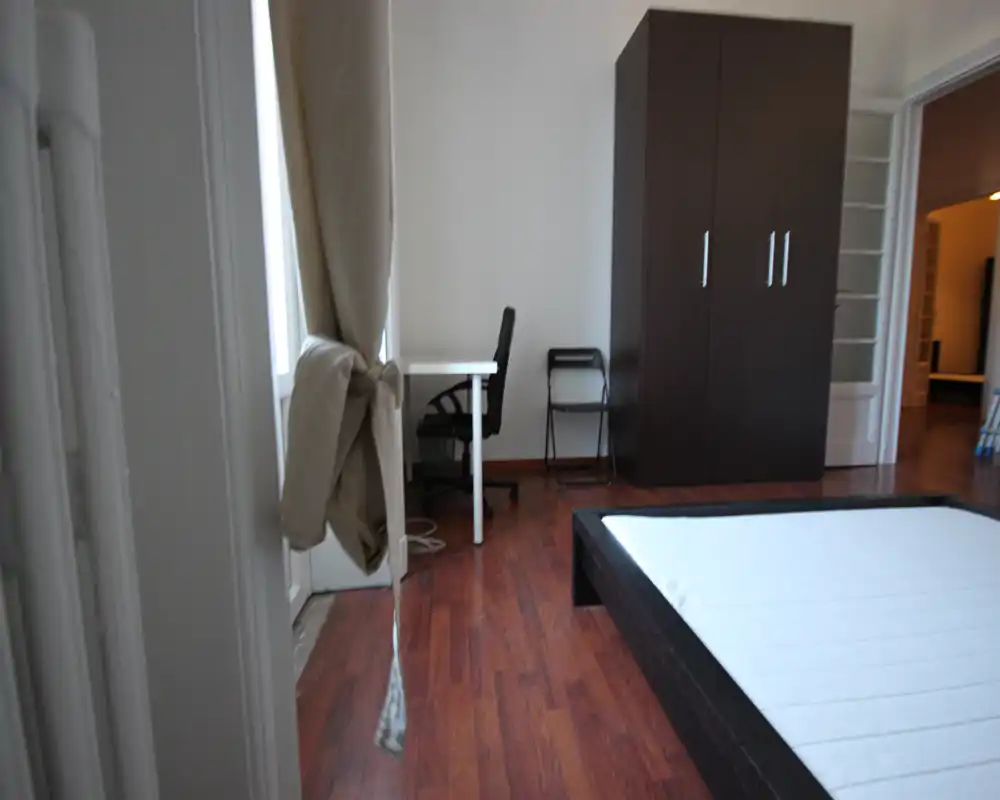 Camera da letto semplice e moderna con letto basso, armadio e una piccola scrivania vicino alla finestra. Luce naturale e pavimento in legno donano un’atmosfera pulita e funzionale, ideale per un annuncio.