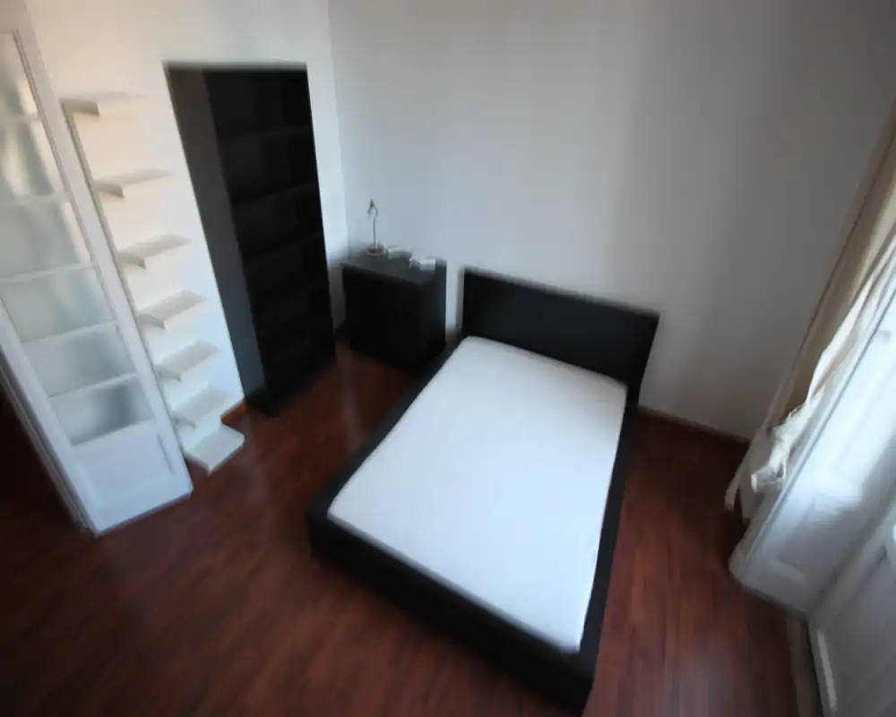 Camera da letto luminosa e arredata in modo semplice con letto matrimoniale, comodino e scaffalatura; pavimenti in legno e ampia finestra con luce naturale.