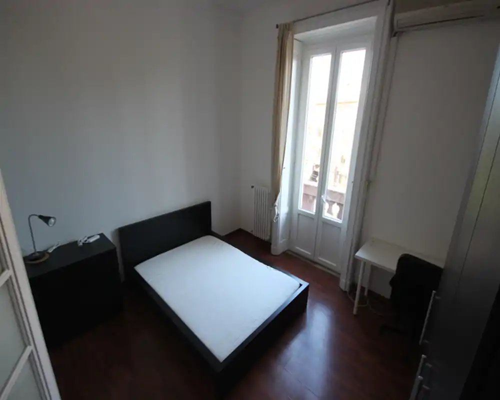 Camera da letto luminosa e minimale con letto matrimoniale, pavimento in parquet e finestra alta che illumina l'ambiente. Scrivania e armadio semplici la rendono pratica per una persona o una coppia.