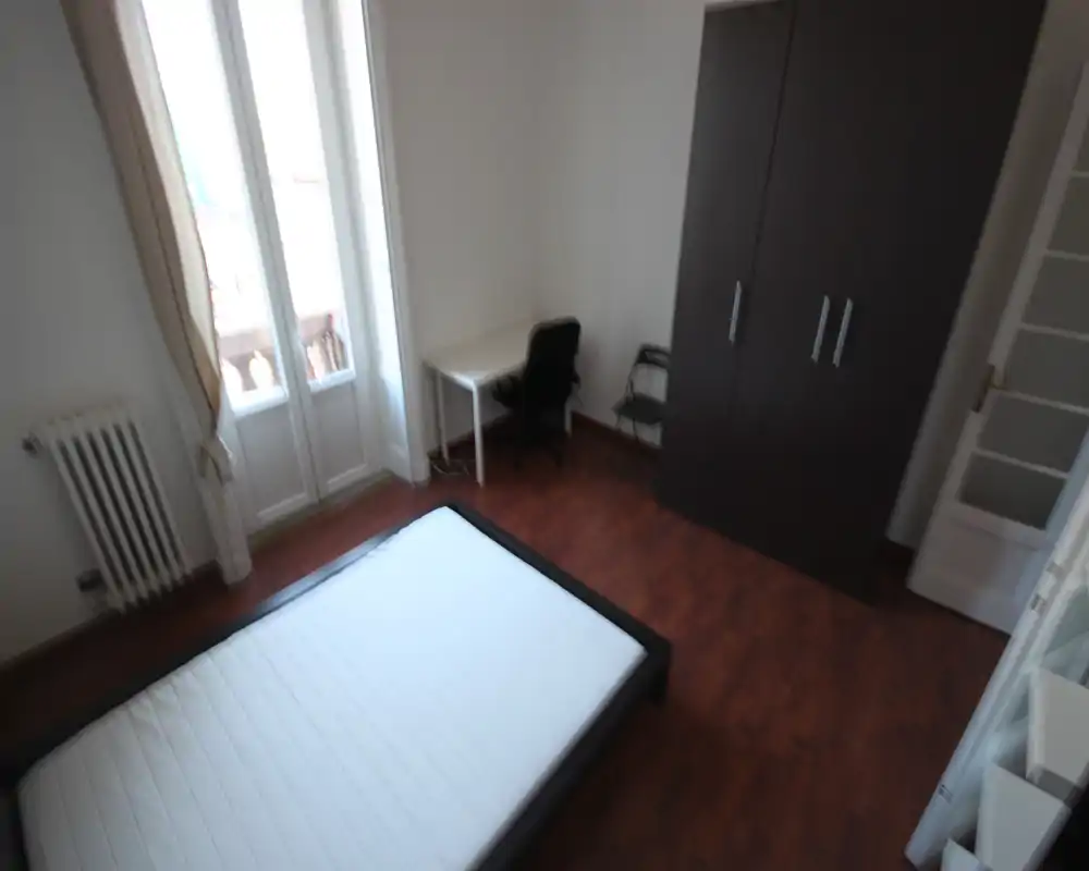Camera da letto luminosa e arredata in modo semplice con un letto matrimoniale, armadio e una piccola scrivania accanto alle alte finestre che garantiscono molta luce naturale.