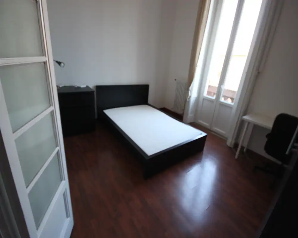 Camera da letto semplice e luminosa con letto matrimoniale su struttura in legno scuro, pavimento in parquet e porta balcone che apporta luce naturale. Arredi minimi come cassettiera e una scrivania creano uno spazio funzionale e tranquillo.