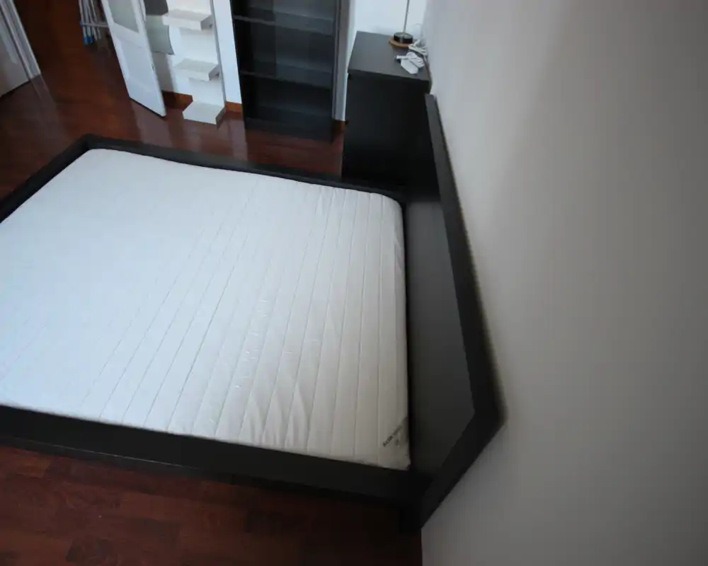 Camera da letto minimalista con un grande materasso su una struttura bassa scura; pavimento in legno e scaffalature a vista. Stile pulito e moderno con buona luce naturale, adatta per annunci di affitto.