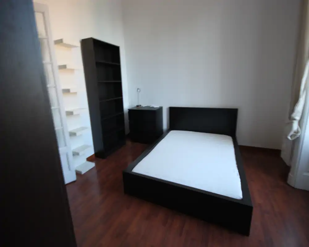 Camera da letto minimal con letto basso nero e materasso, pavimento in legno scuro, scaffali e comodino. Luce naturale a destra che crea un aspetto pulito e moderno, adatto per annunci di affitto.