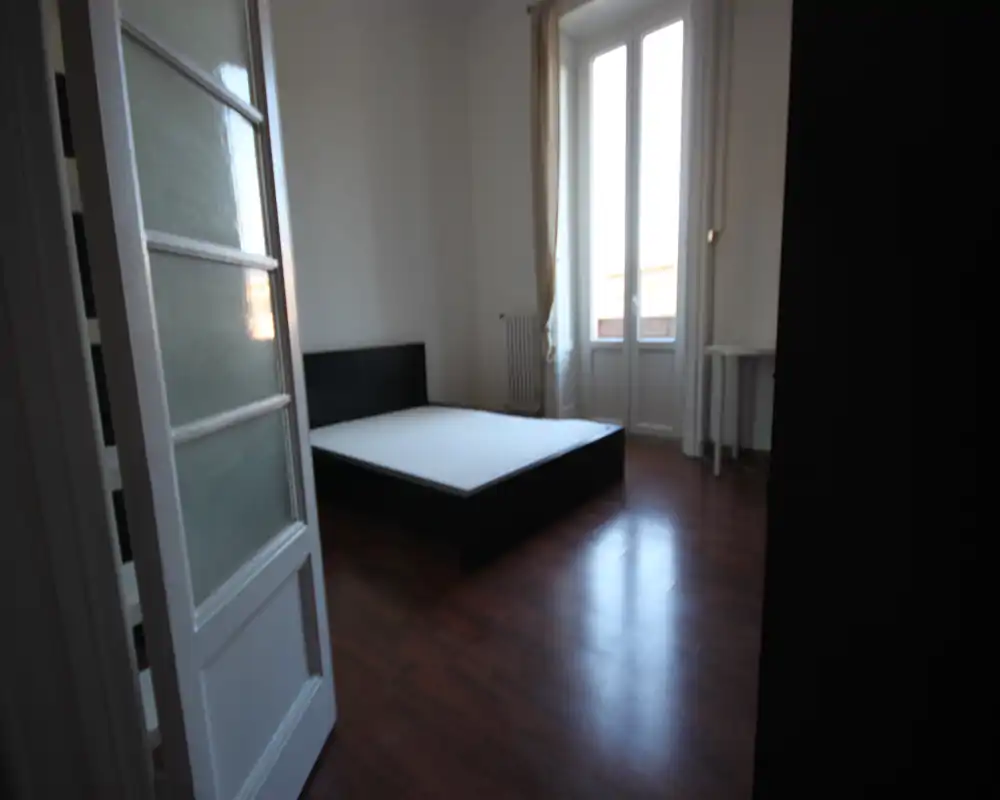 Camera da letto luminosa e minimale con rete matrimoniale e ampia finestra che illumina l'ambiente; spazio semplice, ideale per allestimenti.
