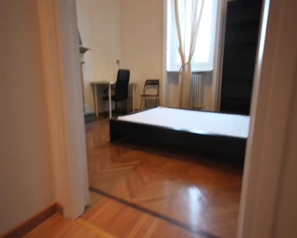 Camera da letto luminosa con struttura letto matrimoniale, pavimento in parquet e una finestra con tende leggere; la stanza dispone anche di una scrivania e scaffalatura, adatta a uno studente o professionista.