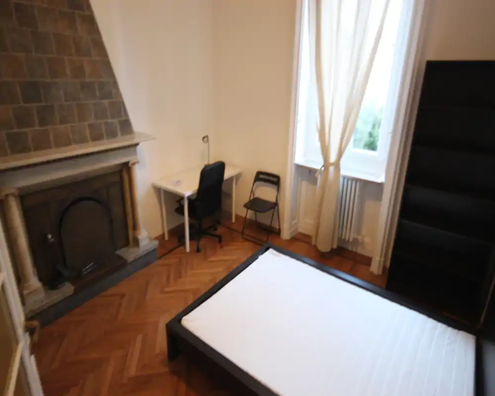 Camera da letto luminosa con struttura del letto matrimoniale, grande finestra con tende, pavimento in parquet e camino decorativo; una scrivania e una libreria completano l'ambiente.