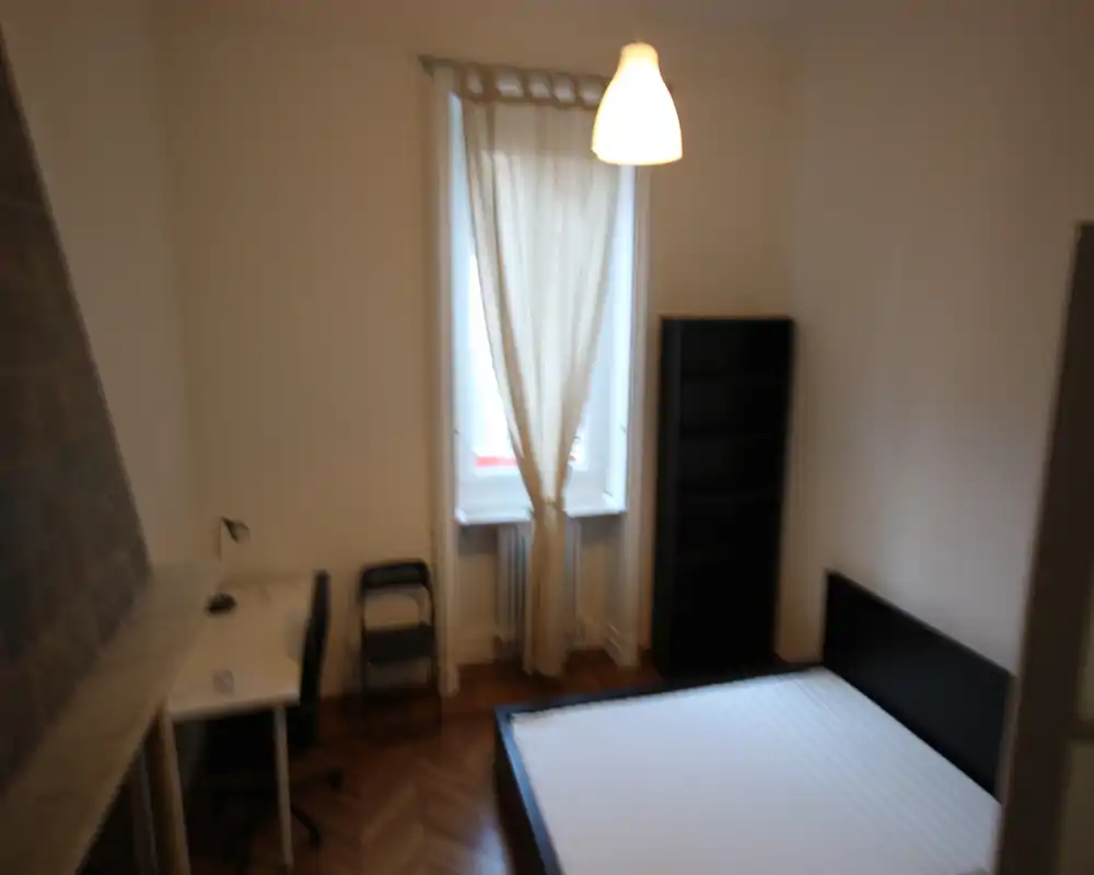 Camera da letto luminosa e arredata in modo essenziale con struttura letto matrimoniale, finestra con tenda e angolo scrivania. Pavimento in parquet e libreria alta, ideale per un annuncio che valorizzi uno spazio notte pulito e minimal.