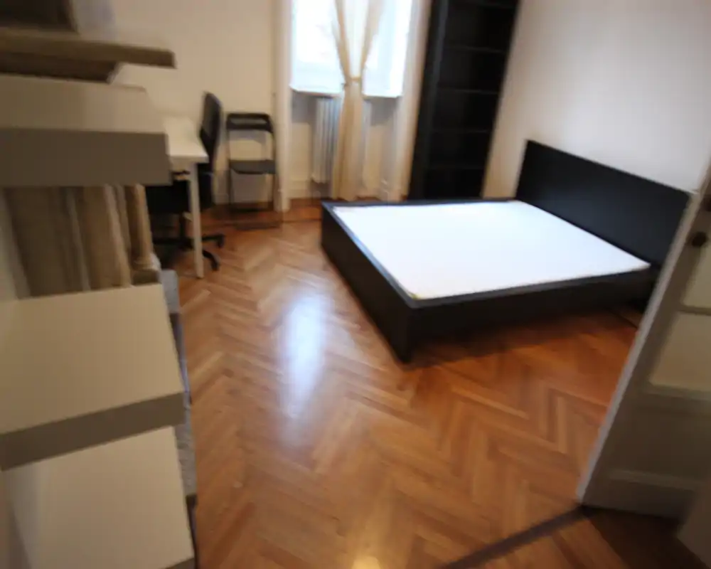 Camera da letto luminosa con letto matrimoniale su struttura nera e materasso, pavimento in parquet a spina di pesce e scaffalatura; luce naturale dalla finestra rende l'ambiente funzionale.