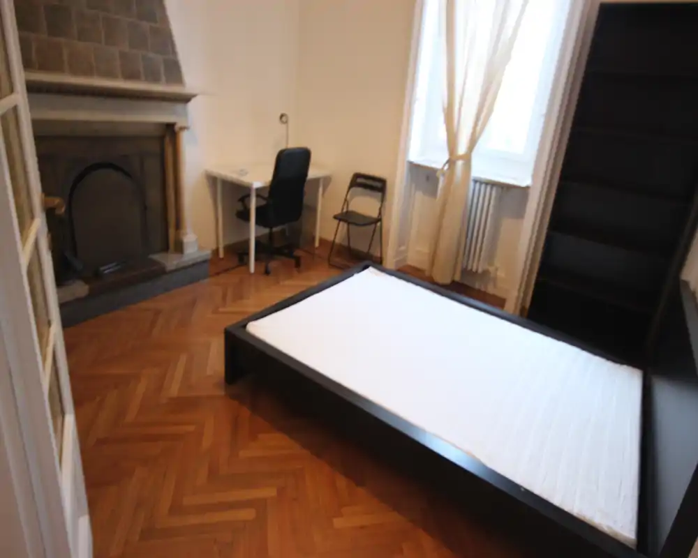 Camera da letto luminosa con letto basso moderno, pavimento in parquet e una grande finestra che offre luce naturale. La stanza è provvista di scrivania, sedia e una libreria alta, adatta per una persona o una coppia.