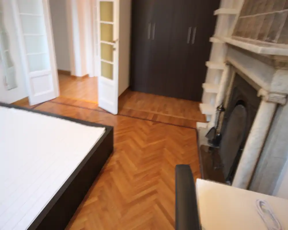 Camera da letto luminosa con struttura del letto visibile, pavimento in parquet e camino decorativo; vista angolata mostra la porta e un armadio adiacente.