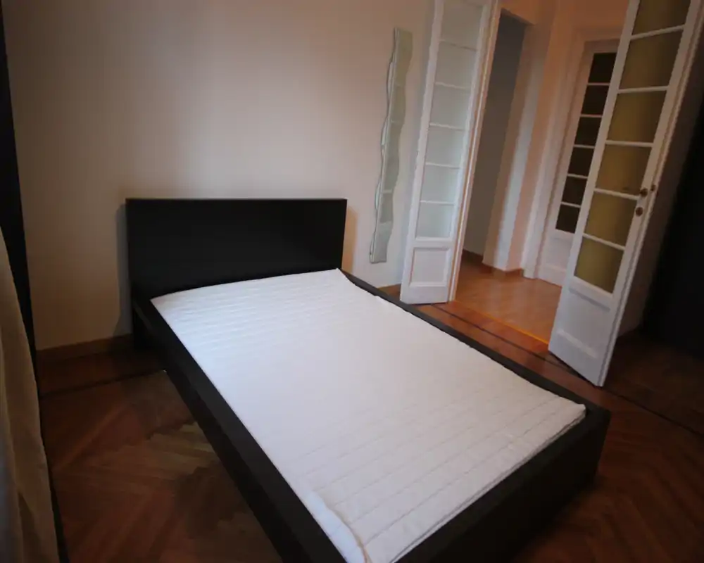 Camera da letto semplice e moderna con letto dalla struttura scura e materasso pulito su pavimento in parquet; porta luminosa e arredi minimi creano un aspetto ordinato.