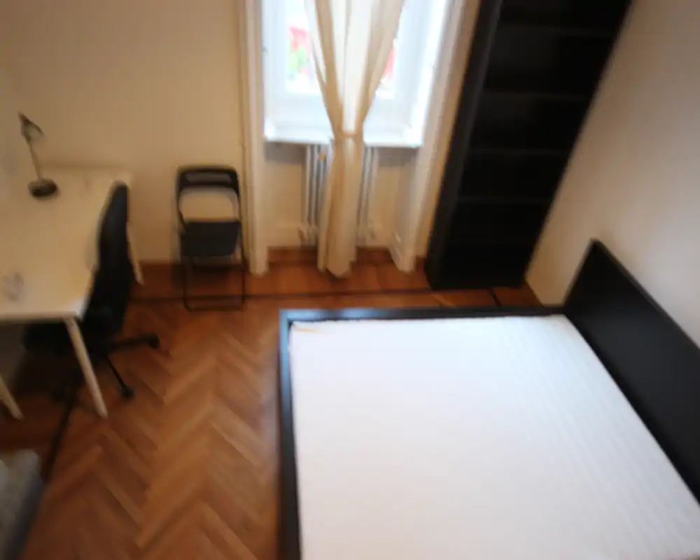 Camera da letto luminosa con un ampio letto visto dall'alto, pavimento in parquet e finestra con tende. Scrivania semplice e scaffalatura rendono lo spazio funzionale e minimale.