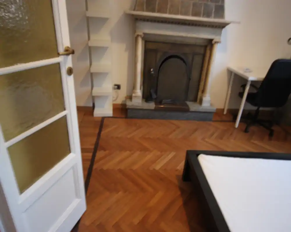 Camera da letto luminosa con parquet, camino decorativo e struttura del letto visibile; ideale per annunci che valorizzano dettagli classici e accoglienti.