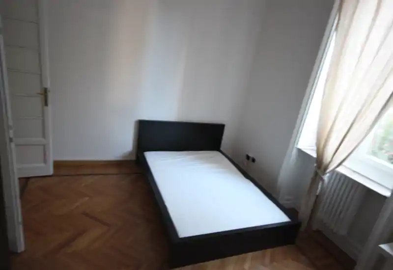 Camera da letto semplice con letto singolo a piattaforma nero su pavimento in parquet, luce naturale intensa da una finestra con tenda e arredamento minimale.
