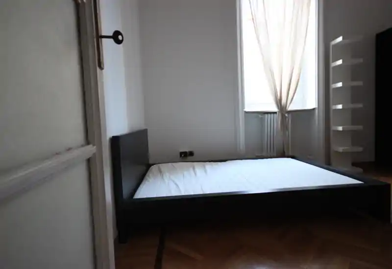Camera da letto minimal e moderna con un letto basso in legno nero e una grande finestra con tende leggere che diffondono luce naturale. Pavimento in parquet e scaffalature semplici per un aspetto pulito e ordinato.