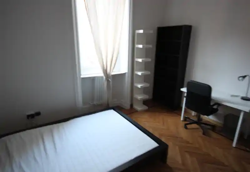 Camera da letto semplice e luminosa con letto matrimoniale, pavimento in parquet, scaffali e una piccola scrivania vicino alla finestra — ambiente minimo e pronto per essere allestito.