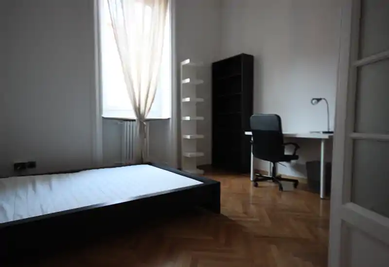 Camera da letto luminosa e minimal con letto basso, ampia finestra con tenda leggera, pavimento in parquet e una piccola scrivania.
