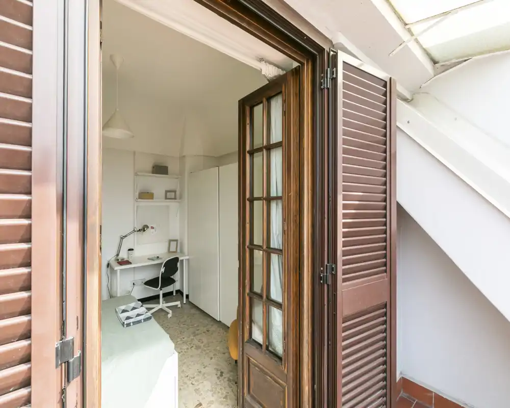 Bright small bedroom seen through wooden shutters, featuring a single bed, desk with chair and shelving — ideal for a student or short-term stay.